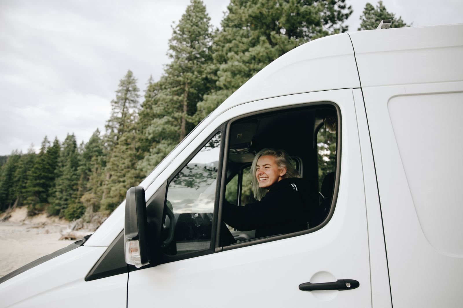 A cheerful woman in a black shirt drives a white van through a scenic forest area.